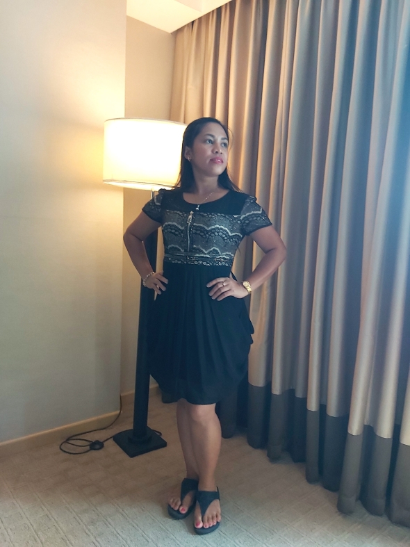 Standing in a refined black dress with a poised posture. A confident and classy moment captured in soft indoor light.